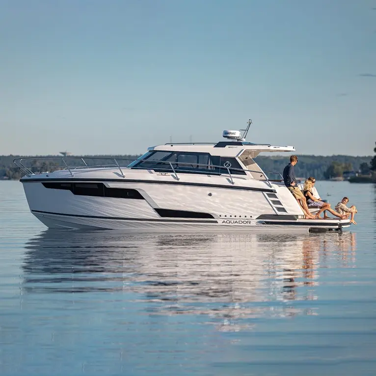 A family enjoys time on an Aquador boat, relaxing on the deck by a calm lake with clear skies and trees in the background.